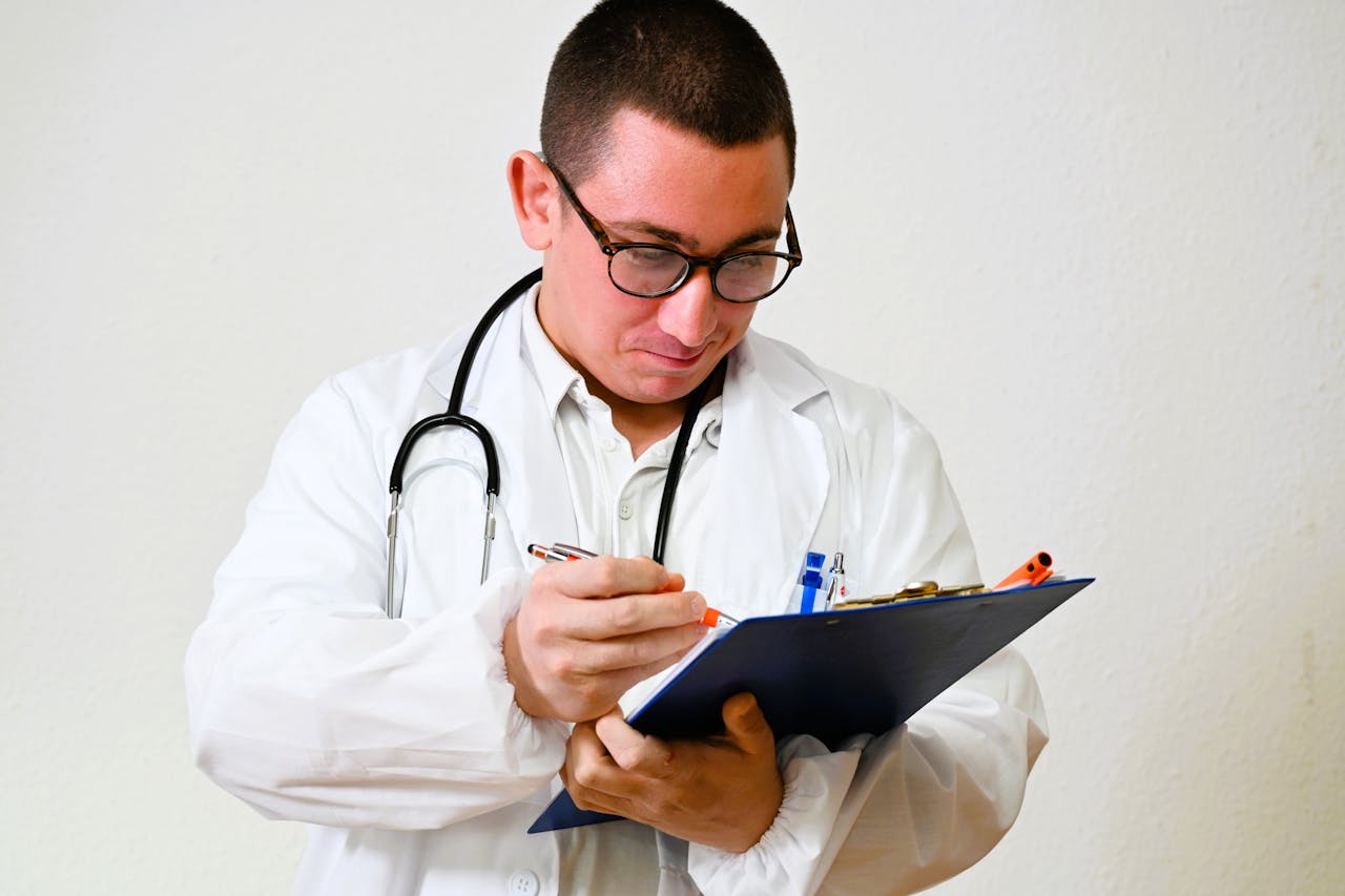 A male doctor in a white coat writes on a clipboard in a hospital setting, showcasing healthcare professionalism.