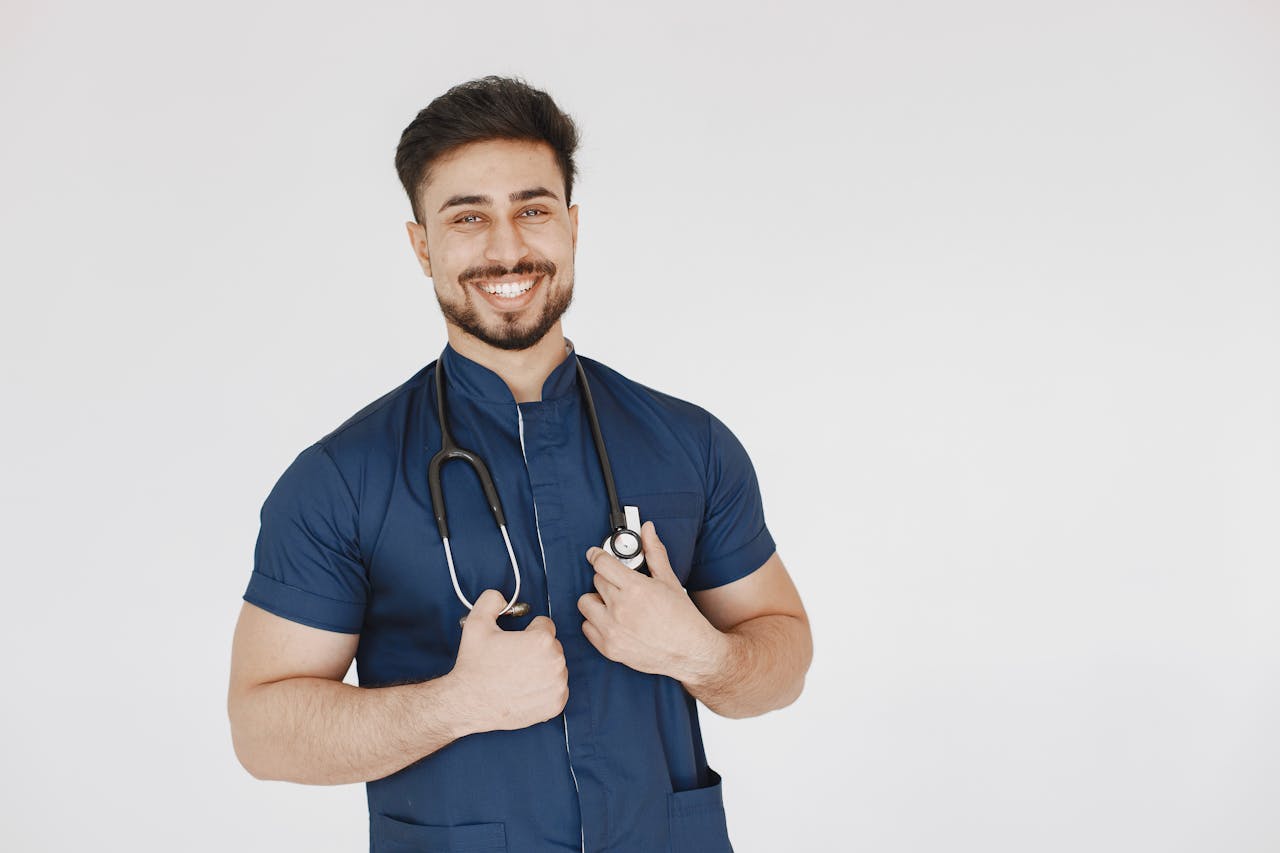 Portrait of a confident male doctor wearing a blue scrub suit and stethoscope, smiling in a studio setting.