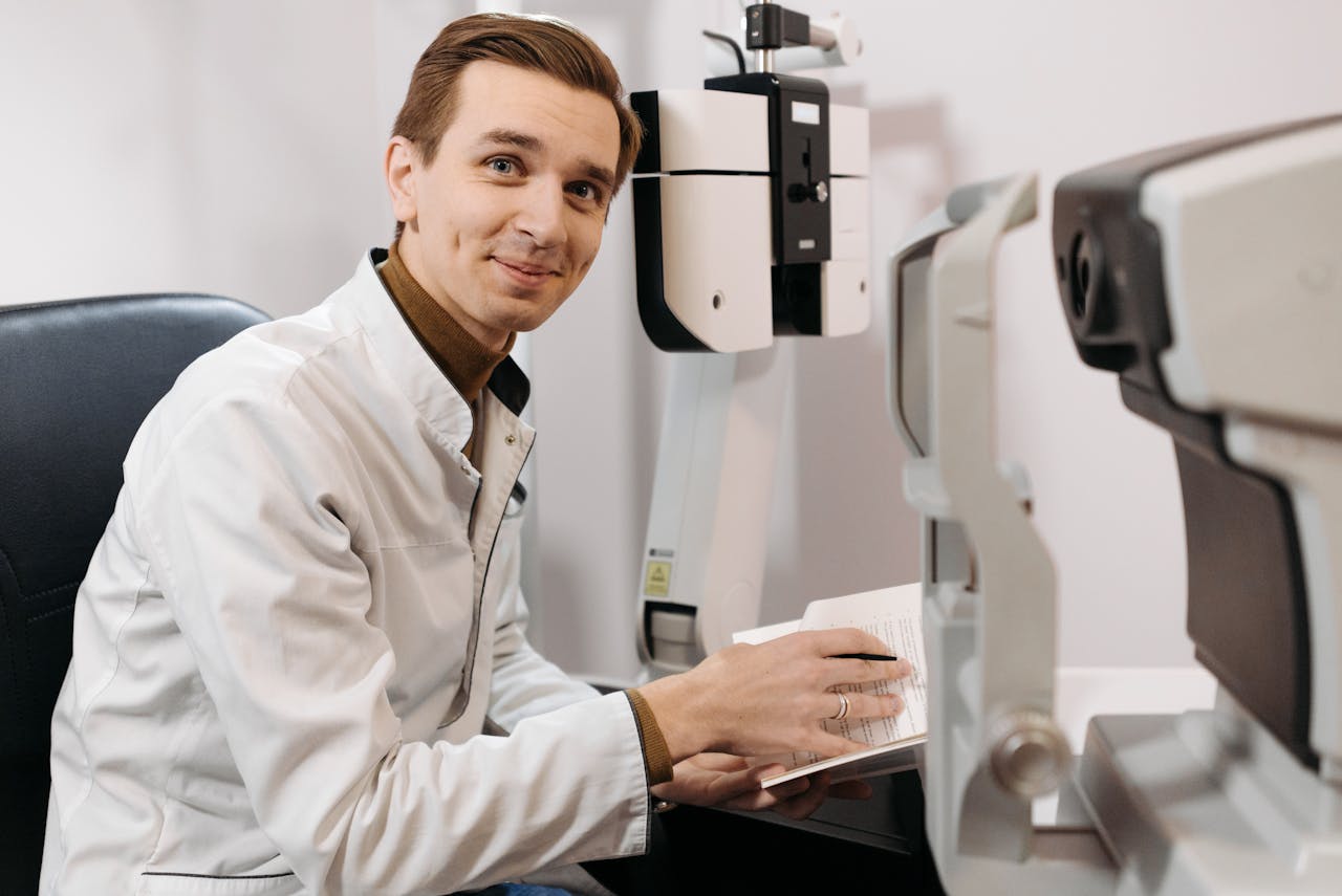 Smiling optometrist wearing a lab coat, taking notes beside eye examination equipment in a clinic.